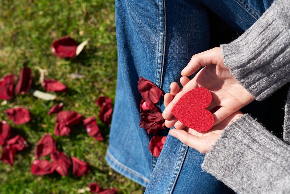 A woman sits on grass holding a red heart-shaped card, surrounded by rose petals, symbolizing love and compassion.