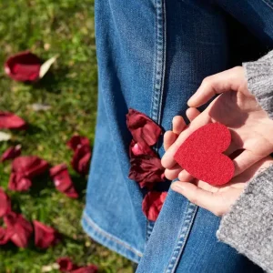 A woman sits on grass holding a red heart-shaped card, surrounded by rose petals, symbolizing love and compassion.